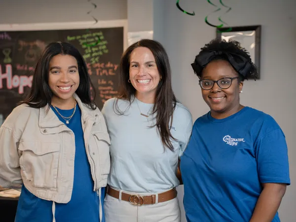 Three smiling female staff members stand together in front of the inspirational Destination Hope chalkboard wall.