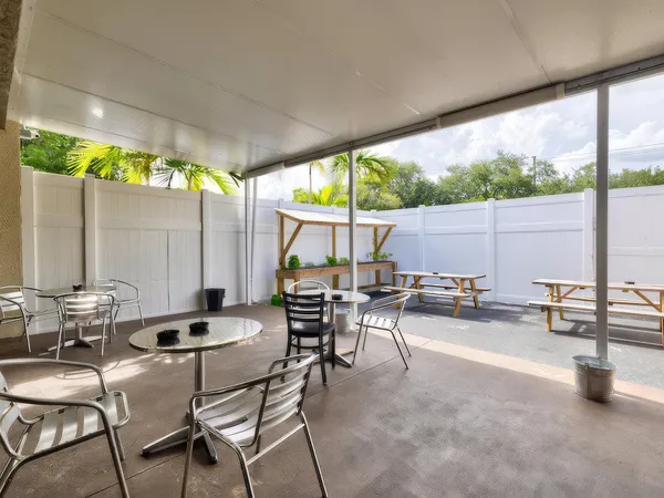 Aluminum tables and chairs stand in a covered outdoor patio area surrounded by white privacy fencing and tall palm trees.