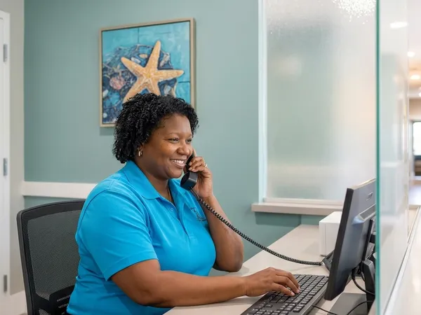 Clinical receptionist smiles warmly while speaking on a telephone receiver and typing at her front desk.