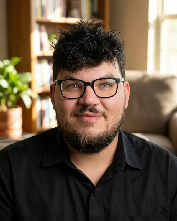 Will, a man in his early 30s with glasses, spiky black hair, and a beard, sits in a sunlit counseling room reflecting recovery.